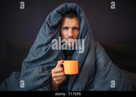 Photo de jeune homme s'assied sur le lit et recouvert d'une couverture bleu foncé. Il est titulaire d'orange tasse de thé. Guy a l'air sur l'appareil-photo. Il est sérieux. Jeune homme est Banque D'Images