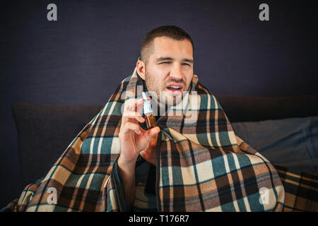 Malade et mal jeune homme détient en pulvérisation nasale mains. Il se rétrécit mais ressemble à la caméra. Guy va éternuer. Il a l'air mauvais. Homme est assis dans la chambre. Banque D'Images