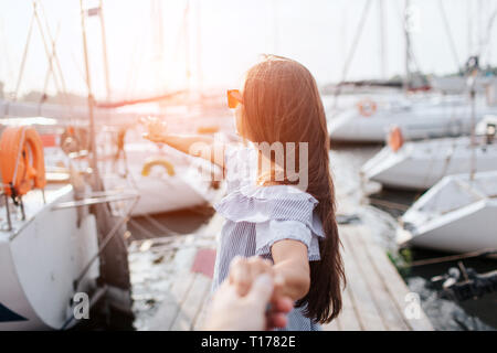 Brune en robe rayée se dresse sur la jetée et de tenir la main. Elle regarde en arrière et le point avec le doigt. Jeune femme porter des lunettes. Il y a beaucoup de voiliers Banque D'Images