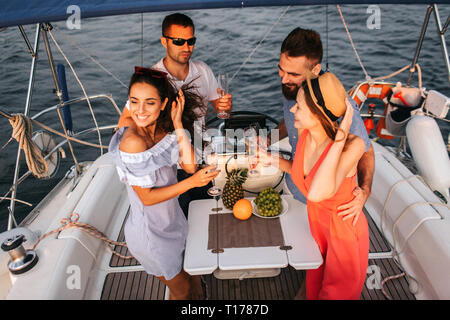 Deux couples sur le yacht de ont partie. Verres de champagne qu'ils contient. Les femmes sont en souriant. Ils ont l'air heureux. Les hommes se tenir avec eux et embrasser leur Banque D'Images