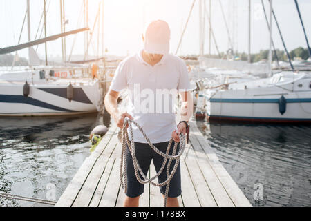 Photo de jeune homme sérieux et concentré. Il se tient sur la jetée et regarder vers le bas. Guy détient des cordes dans les mains. Il y a des bateaux disponibles sur chaque côté du bateau. Banque D'Images