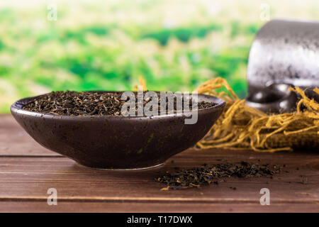 Grand nombre de pièces de thé noir earl grey dans un bol en céramique gris sur tissu du jute avec tasse en céramique noire derrière le champ de thé vert Banque D'Images