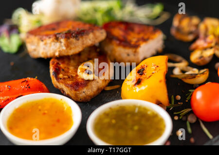 Légumes frais rôti servi avec de tendres médaillons de porc grillé et deux bols de sauce avec l'accent sur la viande Banque D'Images