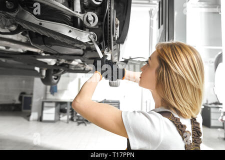 Femme blonde travailler comme mécanicien dans autoservice, réparation des châssis de voiture, à l'aide de la clé. Fille concentré en véhicule fixation autoservice, avec de l'équipement. Levée de l'automobile sur le pont. Banque D'Images