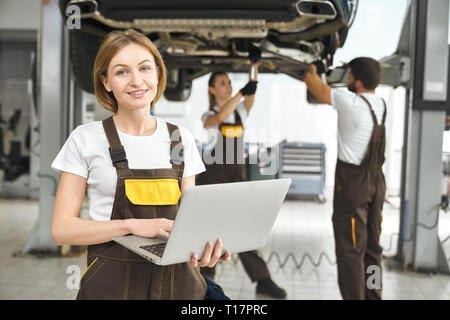 Belle jeune femme, travailleur d'autoservice posing, holding laptop dans les mains. Pretty girl looking at camera, souriant. Fixation de la mécanique automobile, la réparation de pont levé le derrière. Banque D'Images