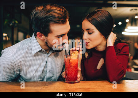 Belle photo de jeune couple assis ensemble et boire un cocktail rouge de la même verre en même temps. Ils ont l'air heureux ensemble Banque D'Images