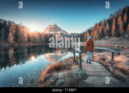 Jeune wooman sur le petit pont de bois contre Lac, forêt d'automne, sommet de montagne, ciel bleu avec du soleil au lever du soleil. Paysage avec girl, réflexion Banque D'Images