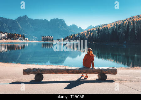 Jeune femme assise sur le banc en bois sur la côte du lac de Misurina au coucher du soleil en automne. Dolomites, Italie. Paysage avec Girl in red jacket, reflec Banque D'Images