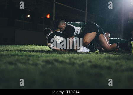 les joueurs de rugby se battent pour le ballon dans un stade de rugby professionnel. Joueurs de l'équipe adversaire qui s'efforcent pour le ballon dans le champ d'herbe pendant le match de nuit. Banque D'Images