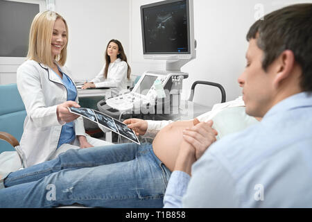 Mari et femme sur l'examen d'ultrasons dans la clinique médicale. Homme tenant la main de femme enceinte, futurs parents à la recherche de x ray. Femme médecin, belle blonde woman showing sonagramme. Banque D'Images