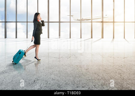 Young Asian woman et valise bleue marche sur le hall de l'aéroport avec fenêtre en verre et flying airplane sur blue sky background Banque D'Images