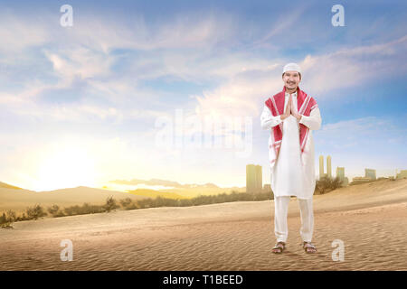 Smiling Asian Muslim man with cap et turban debout et priant le sable avec fond de ciel bleu Banque D'Images