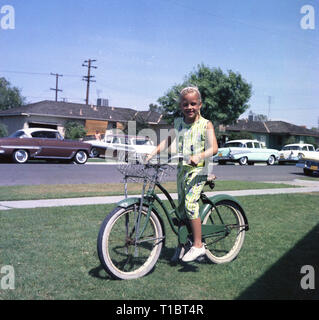 Années 1950, historique, USA, une jeune fille avec son vélo sur un côté herbeux une rue avec des voitures américaines de l'époque sans rouler. Banque D'Images