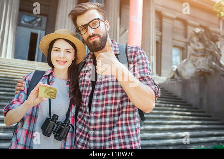 Couple de jeunes touristes stand extérieur à l'escalier et poser sur l'appareil-photo. Elle est titulaire d'une carte gold dans la main. Maintenez l'homme gros pouce vers le haut. Elle a des jumelles sur sa Banque D'Images