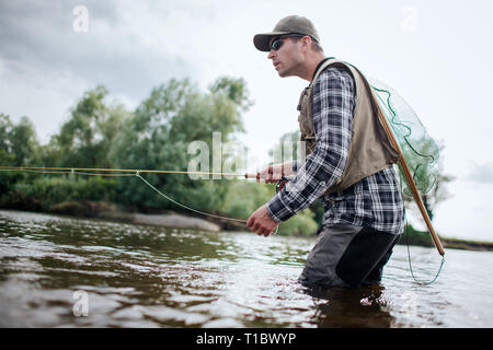 Pêcheur dans l'action. Il se tient dans l'eau et est titulaire d'canne à mouche dans une main et la cuillère dans l'autre. Adultes a également un filet de pêche à l'arrière. Green Banque D'Images