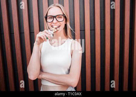 Jeune femme se tient debout à l'intérieur et à la recherche sur l'appareil-photo. Elle est de mordre un morceau de barre de santé. Isolé sur fond rayé. Banque D'Images