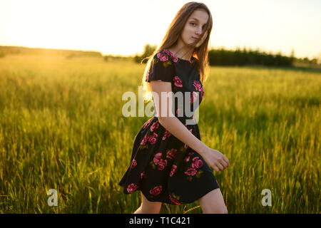 Jeune fille en robe à fleurs adultes, est à pied par Prairie de l'herbe verte. Ses longs cheveux sont fluides sur le vent, toutes éclairées par la lumière du soleil chaud de coucher du soleil. Pays Banque D'Images