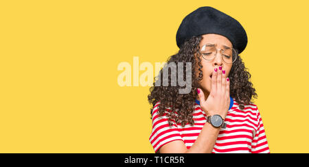 Belle jeune femme aux cheveux bouclés portant des lunettes et de la mode beret ennuyer le bâillement fatigué couvrant la bouche avec la main. Agité et somnolence. Banque D'Images