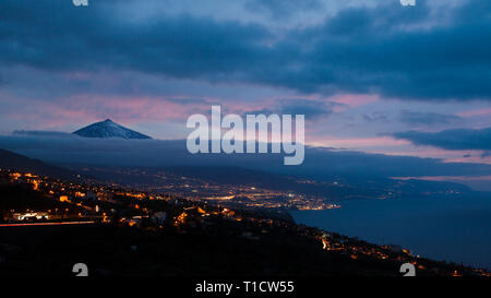Nuages enchanter le Pico del Teide juste après le coucher du soleil avec un magnifique jeu de couleurs violettes et blu. Dans le middleground les lumières de petites villes par t Banque D'Images