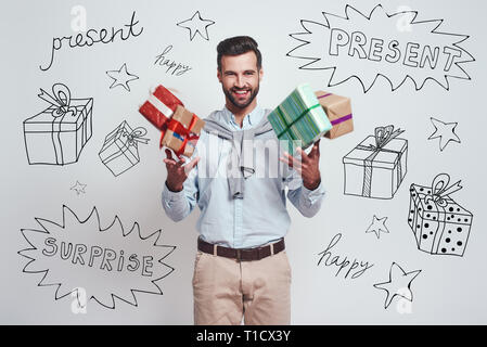 Cadeaux boom ! Cheerful young man holding gifts est et se sentir si heureux alors qu'il se tenait à l'arrière-plan gris avec différentes illustrations doodle sur elle. Banque D'Images