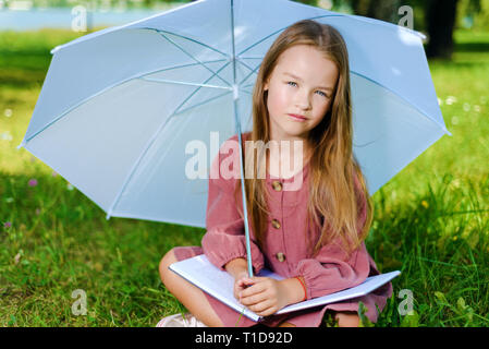 Belle petite fille en robe rose se trouve en position de stationnement sur l'herbe en journée ensoleillée et lit parapluie livre sous Banque D'Images