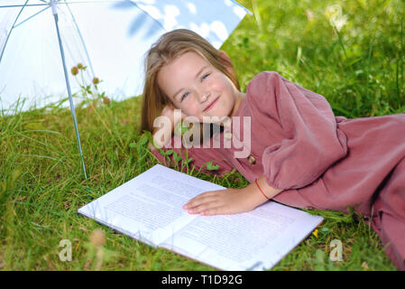 Jolie petite fille se trouve sur l'herbe verte et lit livre sous parapluie blanc in park Banque D'Images