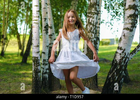 Jolie petite fille en robe blanche et s'enroule et danse entre les arbres du parc Banque D'Images