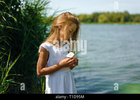 Petite fille romantique pensive en robe blanche près de la rivière tient dans ses mains la fleur Banque D'Images