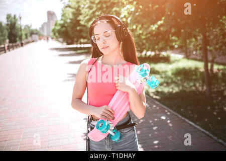 Bon à la fille se tient sur la rue et holding skate dans ses mains. Elle est colorée par des couleurs rose et bleu. Fille est tenue à deux mains et Banque D'Images