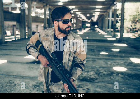 Bel homme sérieux et dans les verres est debout dans le hangar et l'arme dans les mains de paintball holding. Il est à la droite. Homme porte menus Banque D'Images