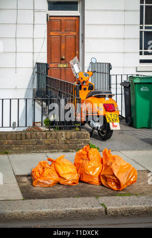 Vue arrière d'un scooter de moteur orange vif à l'extérieur une maison porte avant avec quatre sacs de recyclage d'orange en face de la moto et de la chambre Banque D'Images