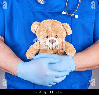 Médecin en uniforme bleu et des gants en latex tenant un ours en peluche brun, fond gris Banque D'Images