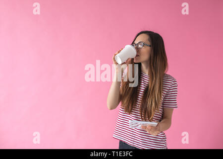 Une belle jeune fille boit du café ou une autre boisson et utilise le comprimé sur un fond rose. Banque D'Images