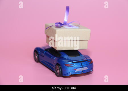 Voiture de sport bleu porte une boîte-cadeau. Le cadeau se trouve sur le toit de la voiture. Fond rose. Le 14 février. Le 8 mars. Petite voiture. Banque D'Images