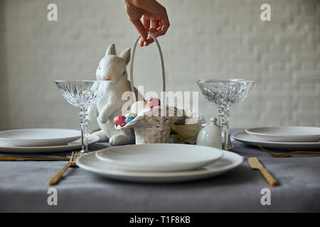 Portrait of woman putting panier avec des œufs peints près de plaques blanches, verres en cristal et bunnie décoratif sur la table à la maison Banque D'Images