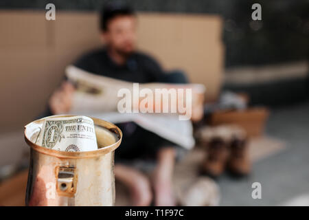 Appareil photo est concentré sur la coupe du métal avec dollar en elle. Il est beggar's cup. Sans-abri est assis sur des cartons et lisant le journal. Il est relaxant. Banque D'Images
