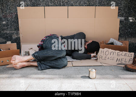 Impuissant et sans défense, l'homme est allongé sur le sol en béton sur carton et dormir. Il est couvrant son visage avec les mains cachant du soleil Banque D'Images