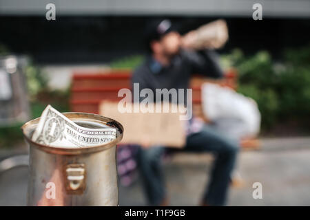 Appareil photo est concentré sur la coupe du métal. Il y a un dollar bill in. Guy est assis sur le banc et l'eau de bouteille. C'est sa tasse. Banque D'Images