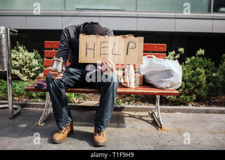 Guy est assis sur un banc et tenant un carton avec de l'aide sur parole. Il a aussi une tasse avec dollar bill in main droite. L'homme est à la mendicité pour sans-abri Banque D'Images