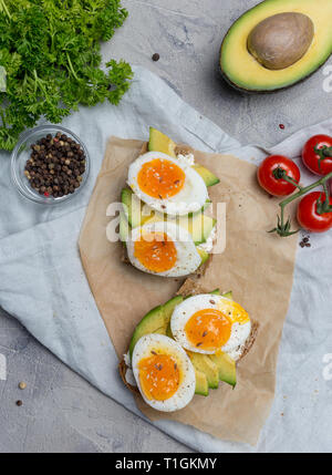 Sandwich sur pain de grains avec oeuf mollet et d'avocat sur le papier kraft, le persil, les tomates cerises, près de serviette sur fond gris. Concept d'alimentation saine. Banque D'Images