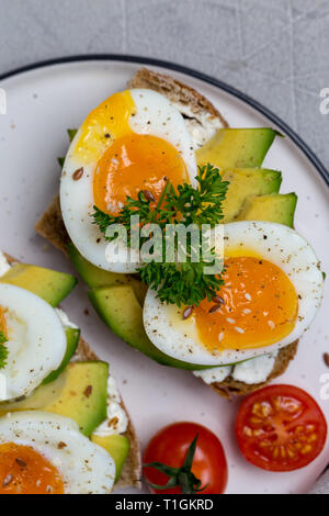 Sandwich sur pain de grains avec oeuf mollet et avocat sur plaque blanche, persil, tomates cerises, près de l'avocat sur fond gris. Concept de santé Macro Banque D'Images