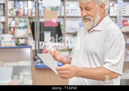 Cheerful pensionné holding prescription papier et bouteille en plastique blanc avec des pilules. Les chimistes de magasin, le choix des médicaments à partir de la maladie. Vieux barbu ayant les cheveux gris. Banque D'Images