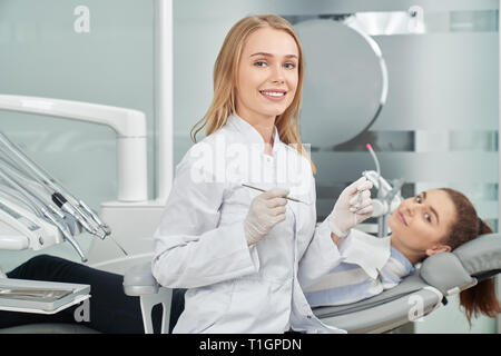 Charmante femme dentiste posing. Beautiful woman smiling at camera, à la restauration et la tenue d'instruments. Derrière le client sur l'art dentaire président. Happy woman en blouse blanche. Banque D'Images