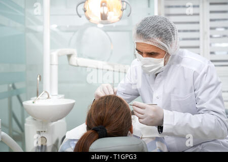 Dentiste dents traitement de client en clinique dentaire privée. Stomatologist portant en sarrau blanc, masque médical, le chapeau et gants. Lampe, lavabo blanc et dentaire au cabinet. Banque D'Images