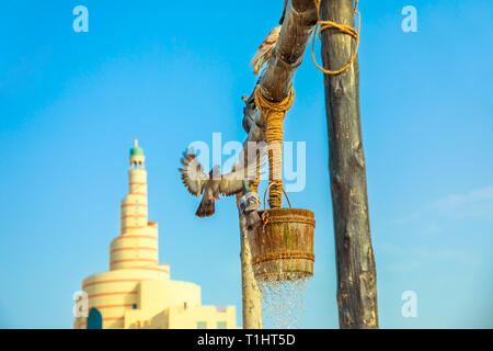 Pigeon vole à vieux puits fontaine, célèbre lieu touristique au milieu de Souq Waqif en face de la mosquée et minaret dans centre de Doha, au Qatar. Moyen Orient Banque D'Images