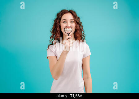 Jolie fille brune avec les cheveux bouclés sur fond turquoise bleu isolé avec une loupe montrant ses dents chez le dentiste. Stomatologie Banque D'Images