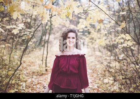 Belle jeune fille avec des cheveux noirs bouclés dans un haut marron dans un parc en automne, smiling at the camera Banque D'Images