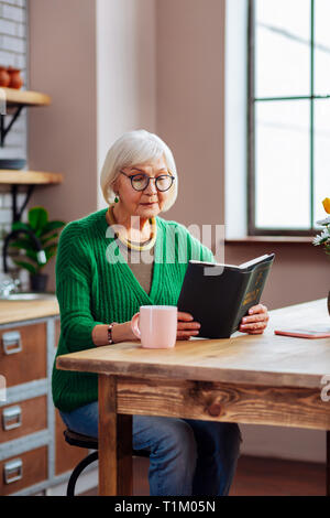 Dame septuagénaire assis à table de cuisine avec Bible en mains Banque D'Images