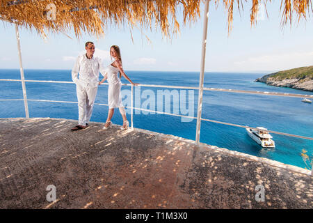 Vue romantique du couple posant dans des vêtements blancs, debout sur un balcon extérieur avec beau paysage de mer grecque bleue. Concept de l'amour de la lune de miel Banque D'Images