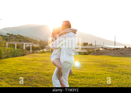 Vue romantique de l'heureux couple en habits blancs, lorsqu'ils caresser sur l'herbe verte. Magnifique paysage de montagne au-dessus de soleil au coucher du soleil. Concept d'h Banque D'Images